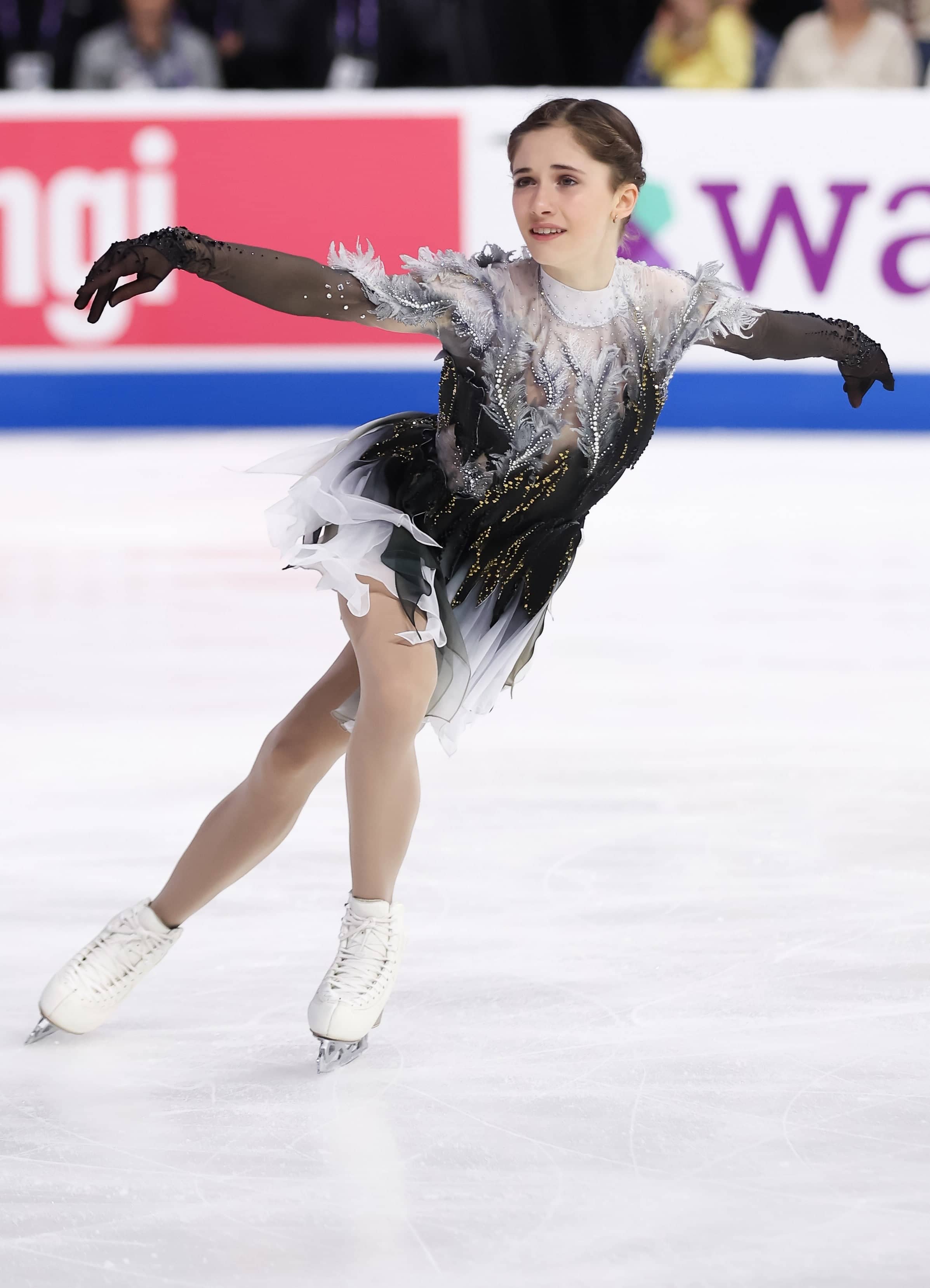 Isabeau Levito skates with her arms outstretched. She is a young woman with brown hair tied back in braids. She is wearing a black skating costume with grey and white feather details and long sheer black gloves